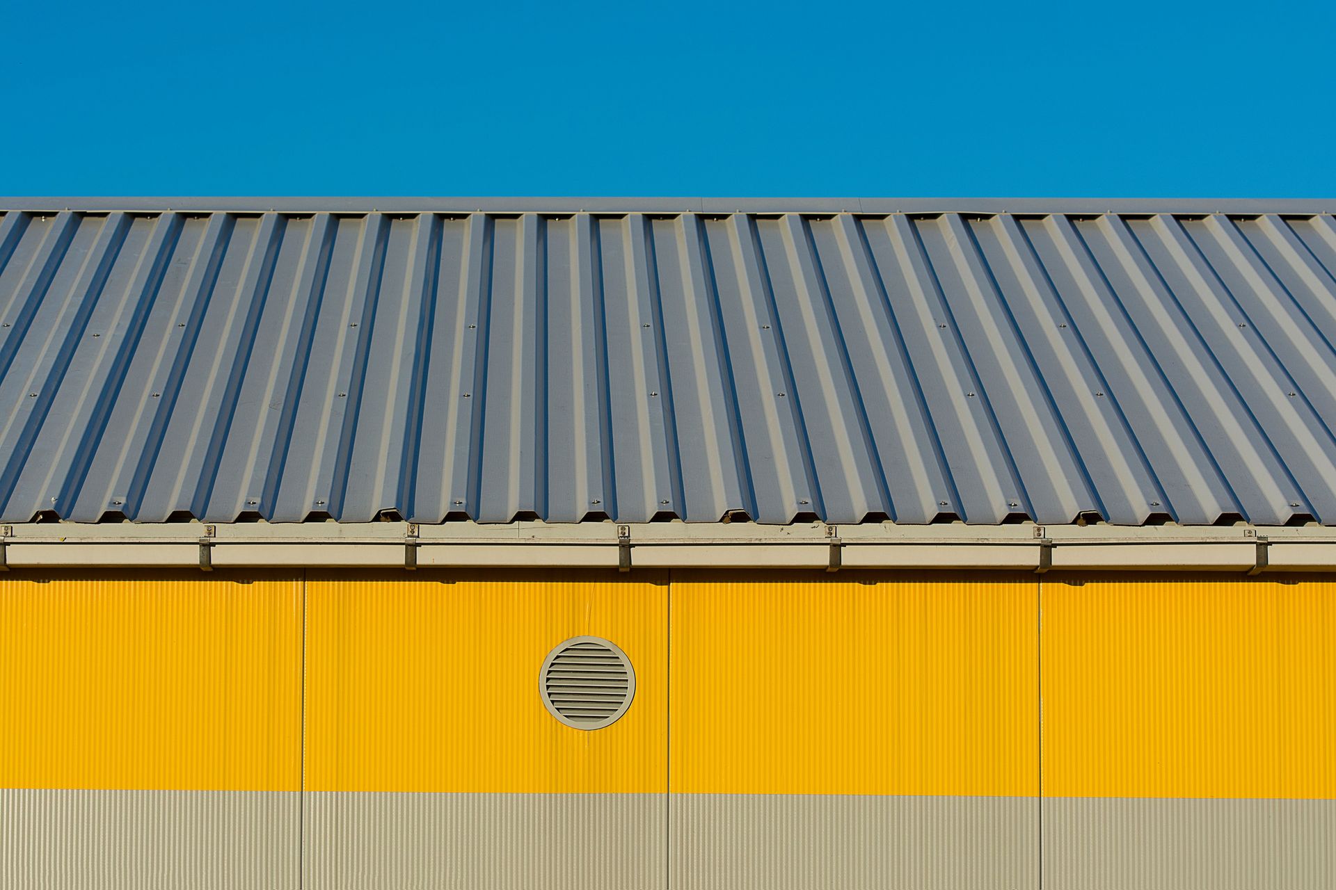 Modern industrial building's yellow facade with metal roof against clear blue sky.